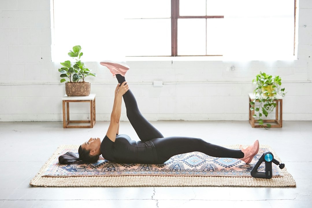 Woman stretches hamstring on patterned rug with recovery tool.